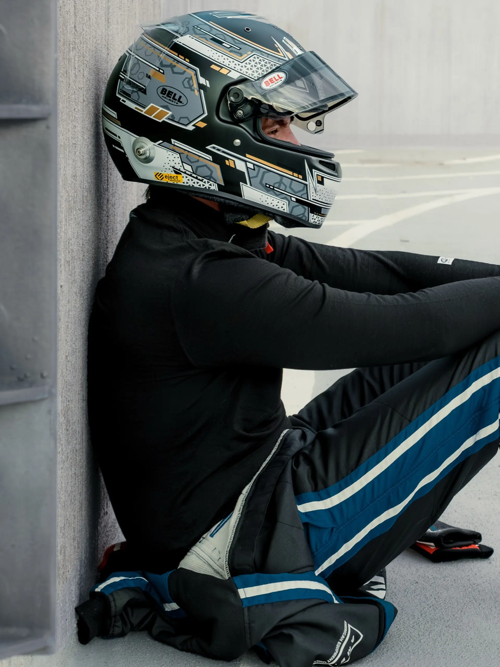 Randy Kinne in his Bell helmet and race suit, visor up, sitting against a wall between sessions.