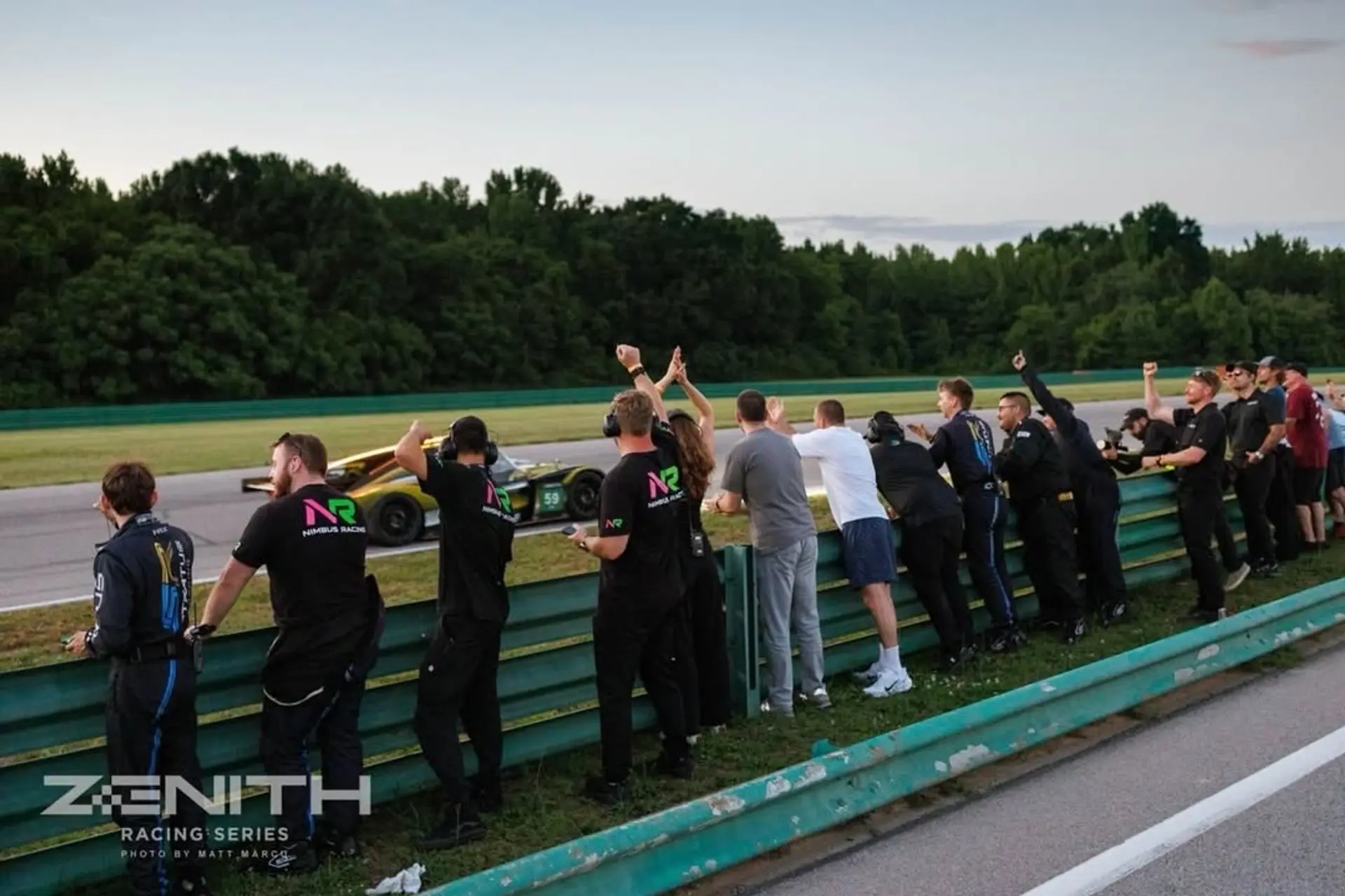 Crew leaning over the pit wall at dusk at VIR as the #59 comes past on track.