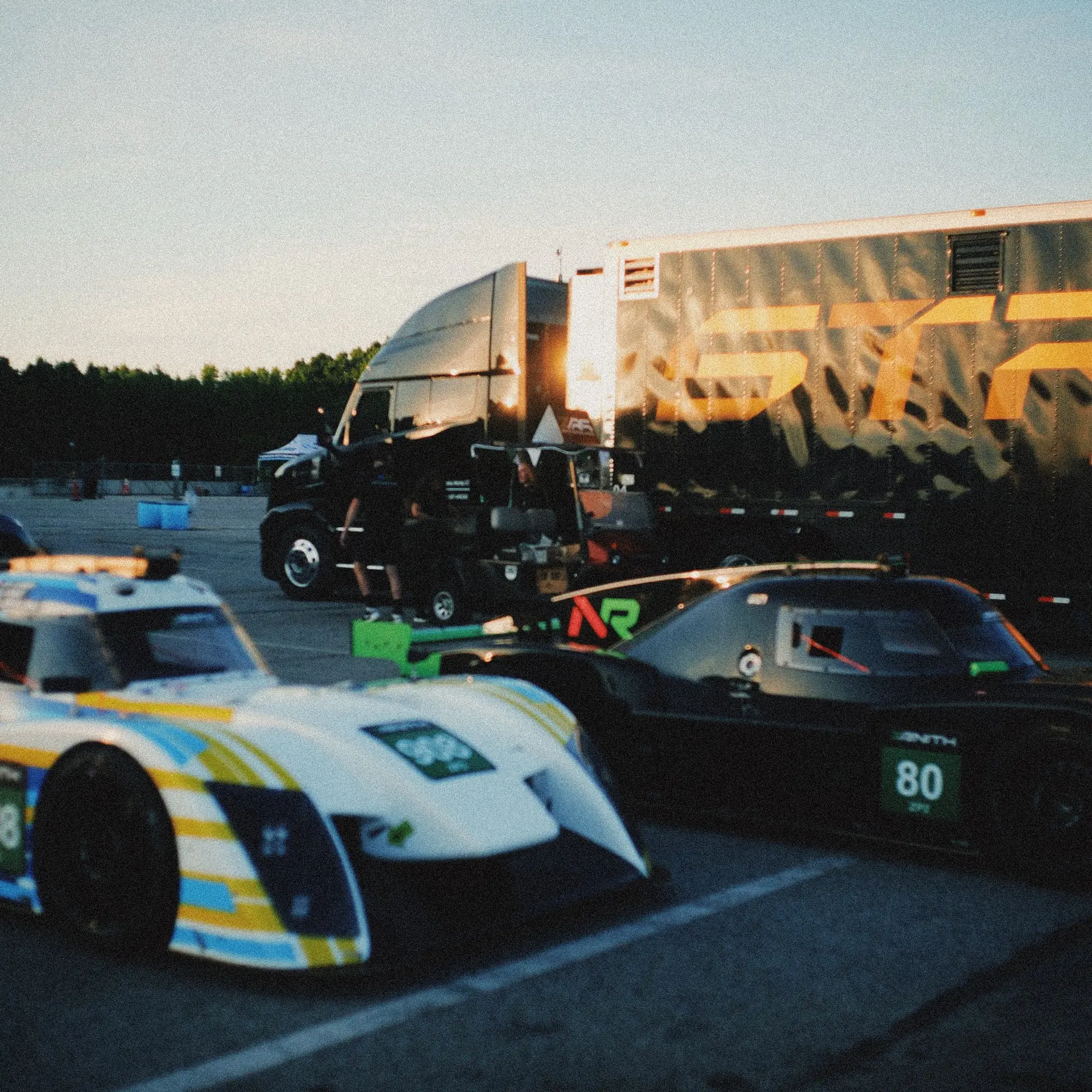 Prototypes parked in the VIR paddock at dusk, a team's transporter behind them in warm low light.