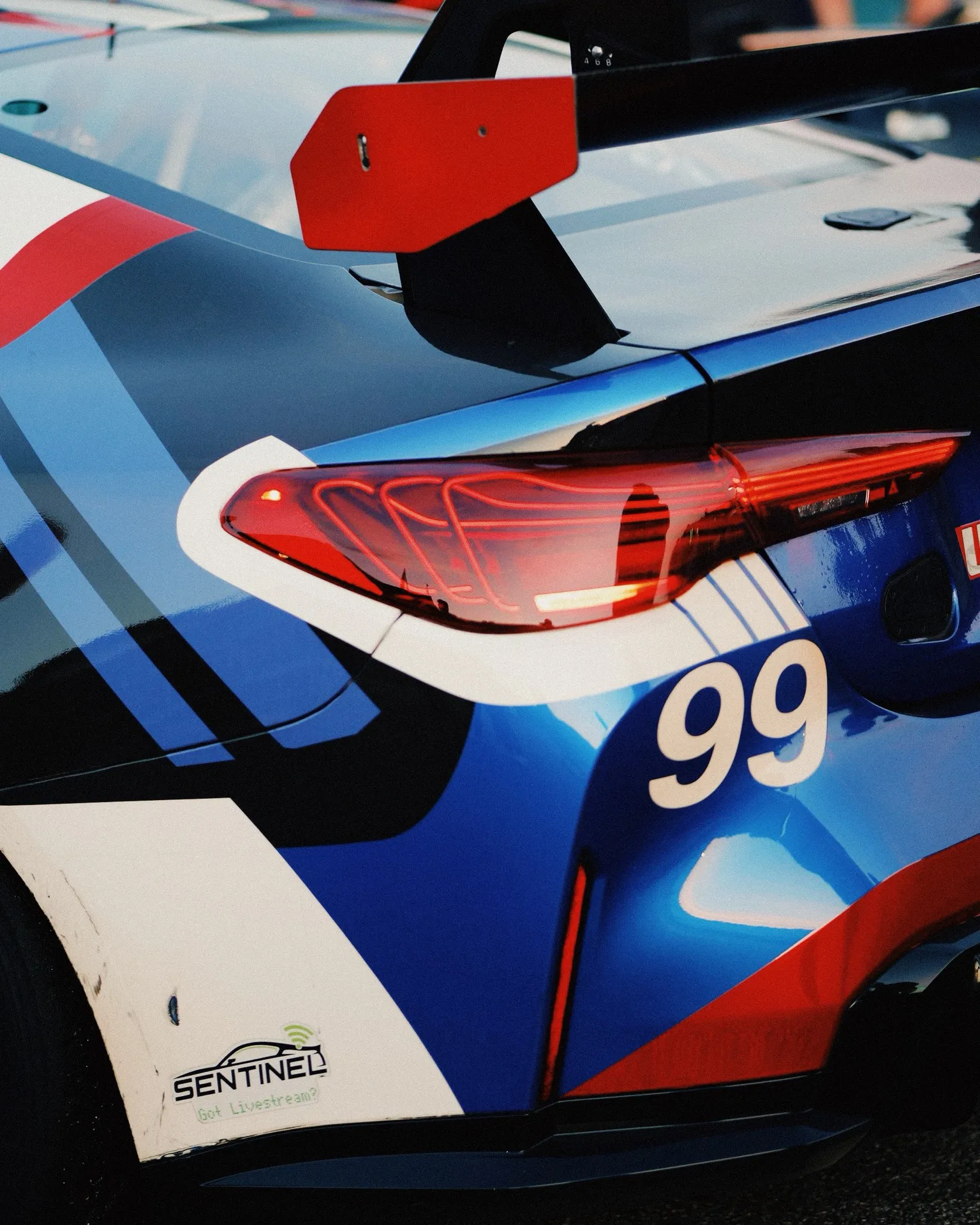 A close detail of the #99 BMW M4 GT4's rear quarter, the number 99 and tail light against the blue, red and white livery.