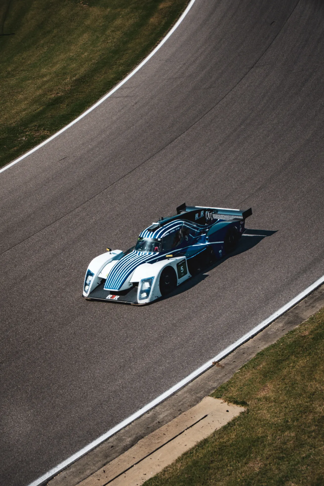 Overhead view of the #5 prototype cutting through a downhill corner, its shadow stretched across the tarmac.