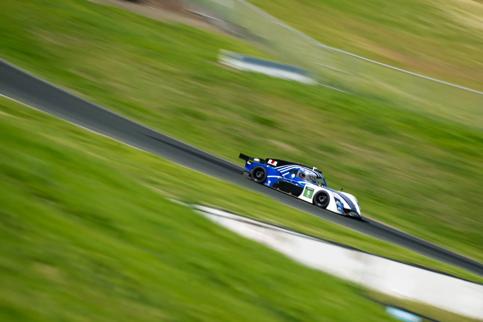 The blue-and-white Round 3 Racing #5 prototype at full speed, panned against the grass banking.