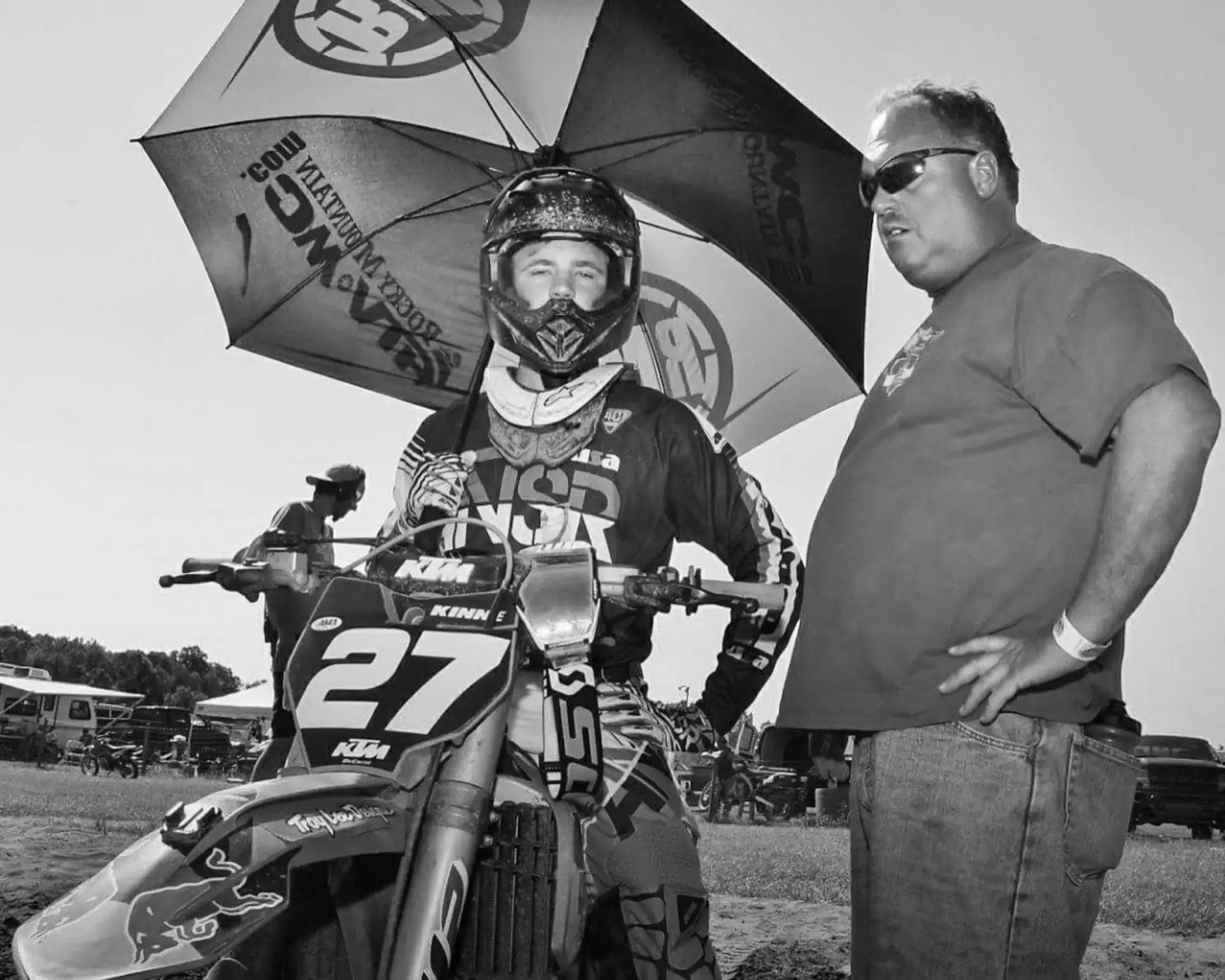 A young Randy Kinne in motocross gear on his #27 KTM under an umbrella, his father standing beside him at the track.
