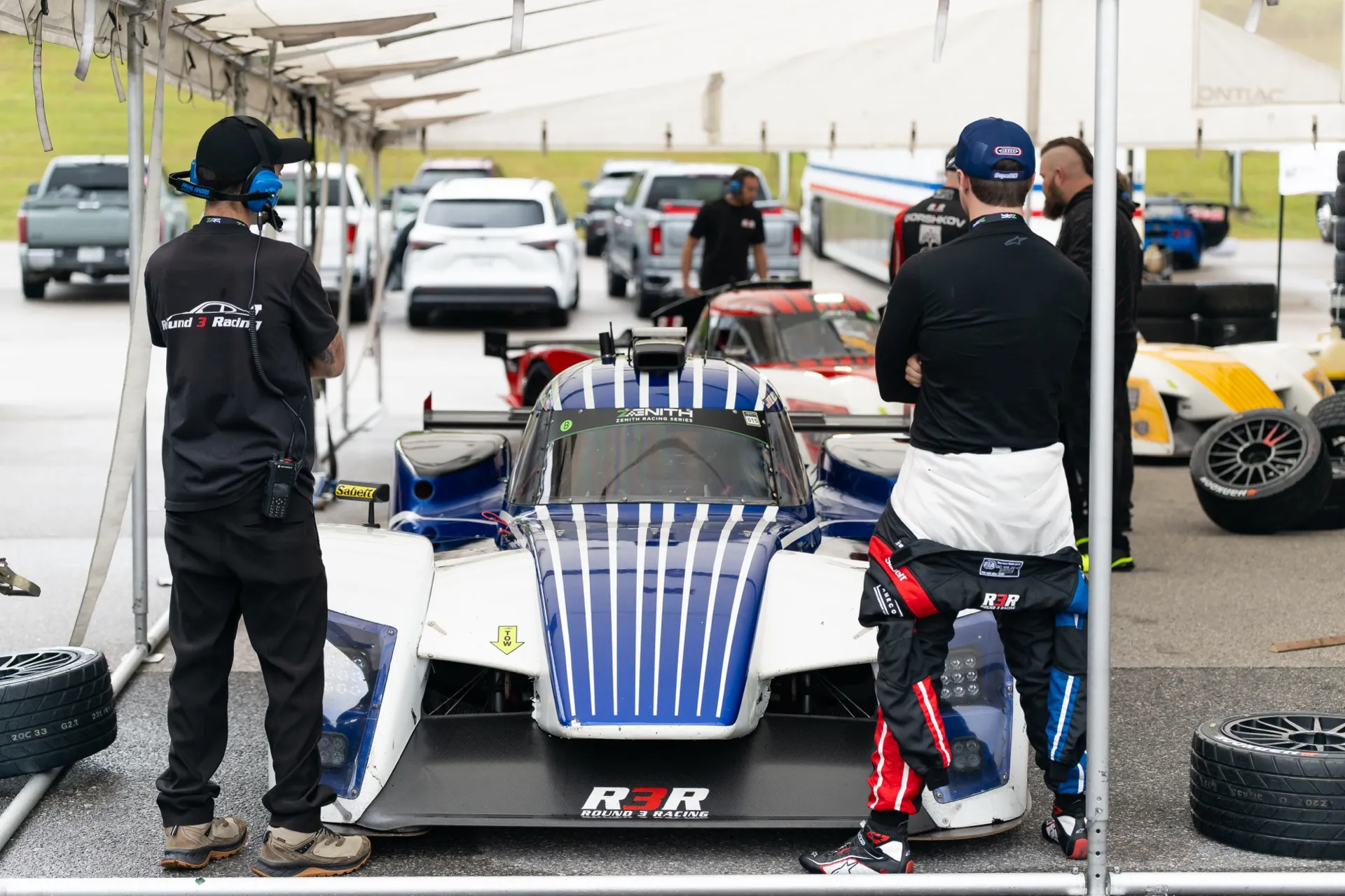 The blue-and-white Round 3 Racing #5 prototype under the team awning in the paddock, two crew members working around it.