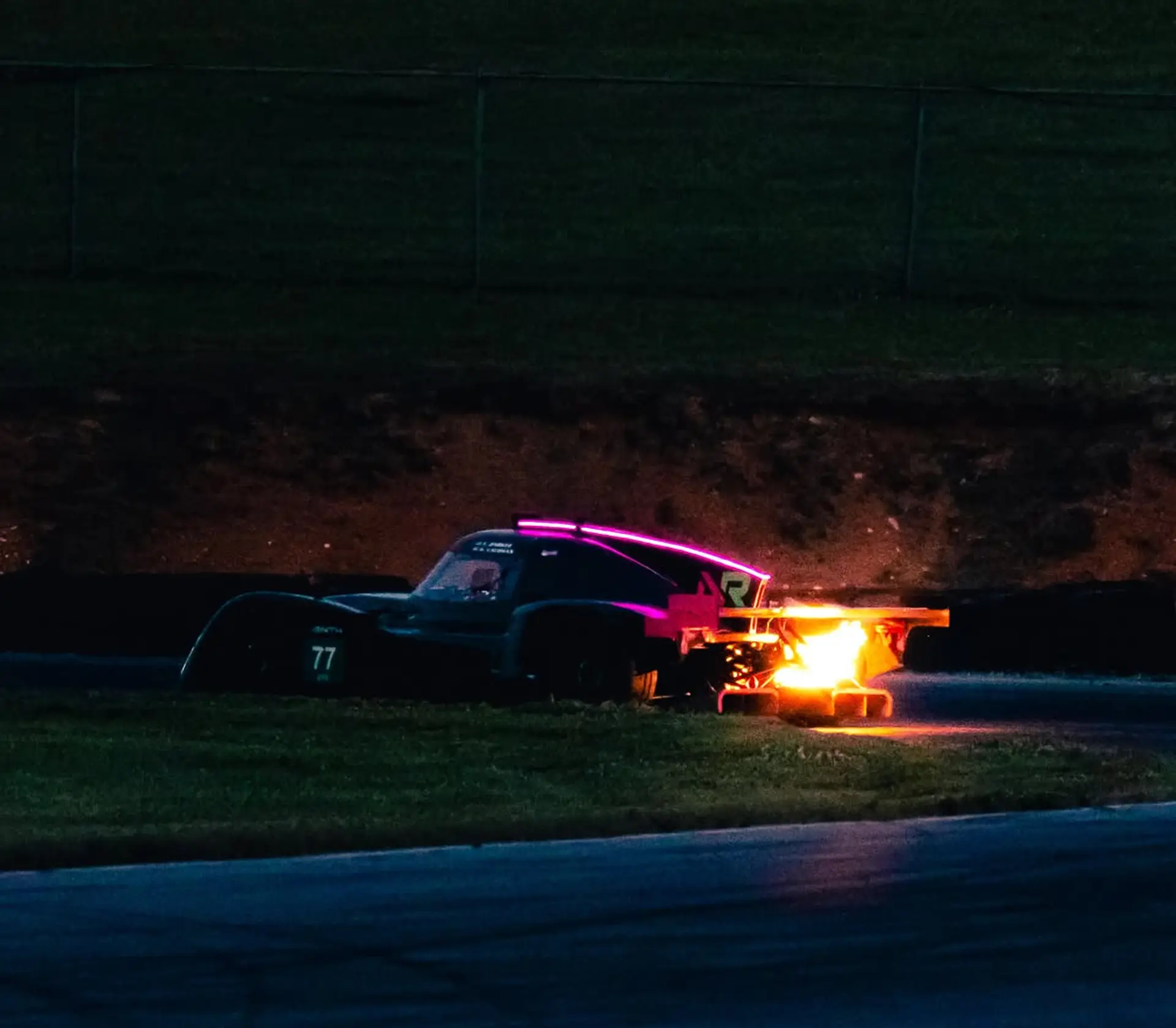 The Nimbus #77 at night at VIR, a burst of flame from the side exhaust lighting the dark banking, the car's pink light strip glowing along the top of the bodywork.