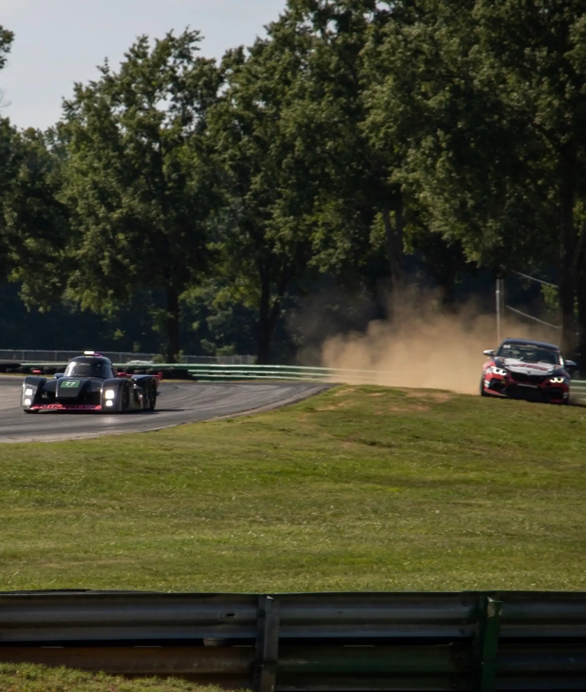 The Nimbus #77 prototype at speed at VIR, a GT car kicking up a cloud of dust off-track in the background.