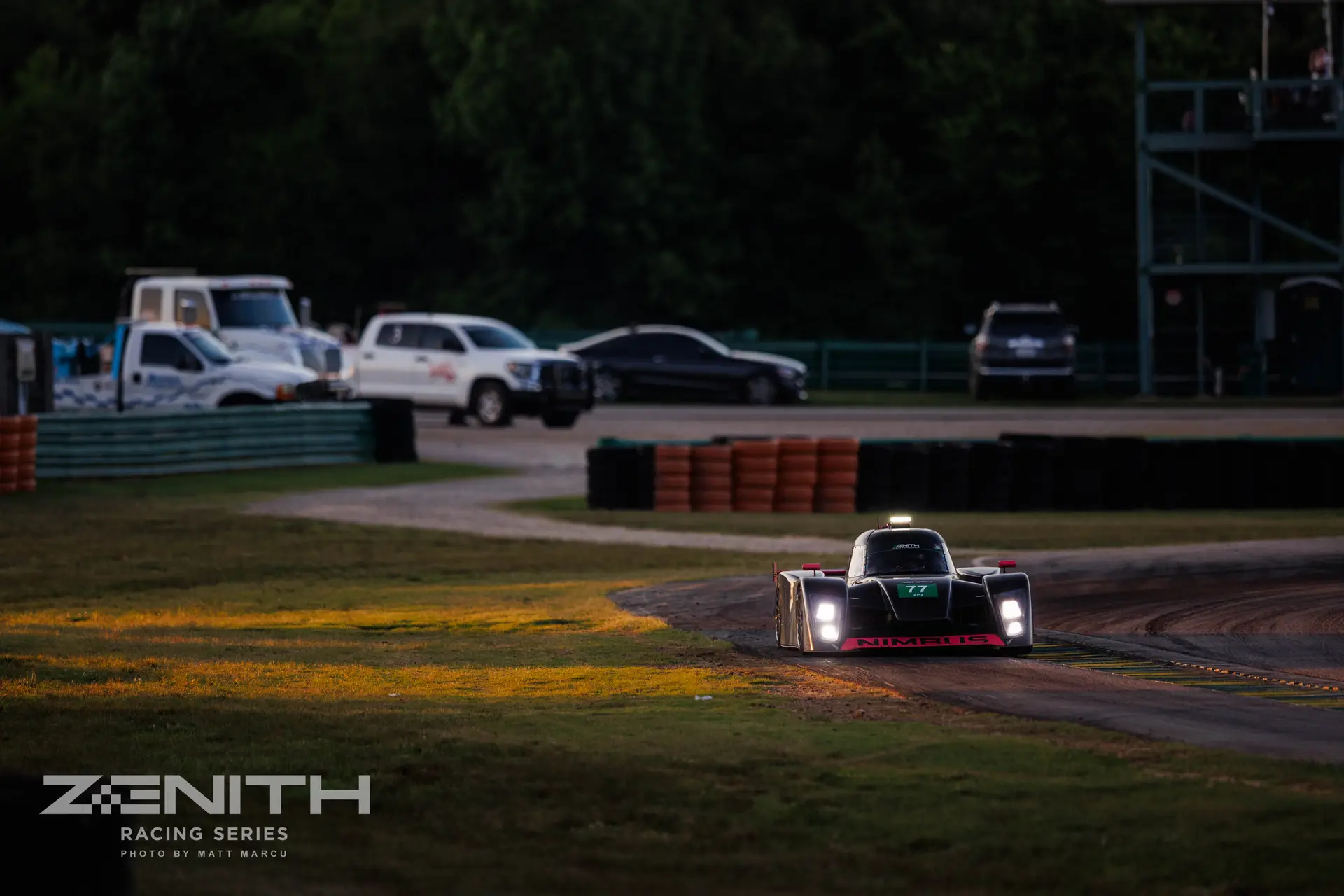 The Nimbus #77 at dusk at VIR, headlights lit, crossing a golden-lit stretch of grass with the dark tree line behind.