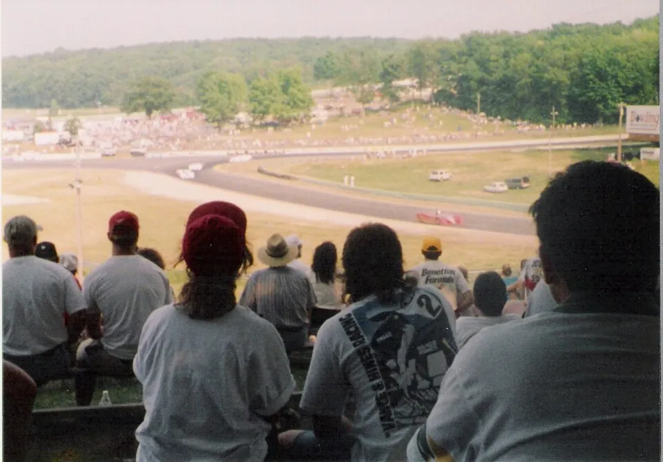 Spectators on a grassy hillside watching cars sweep through a corner, an older photograph.