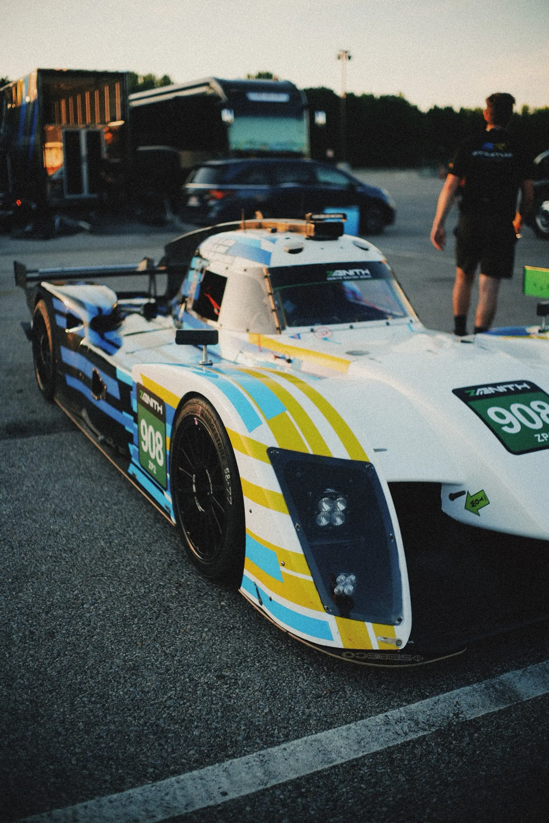 Automatic Racing's white, blue and yellow NP01 prototype, car 908, in the VIR paddock at dusk with a crew member walking past.