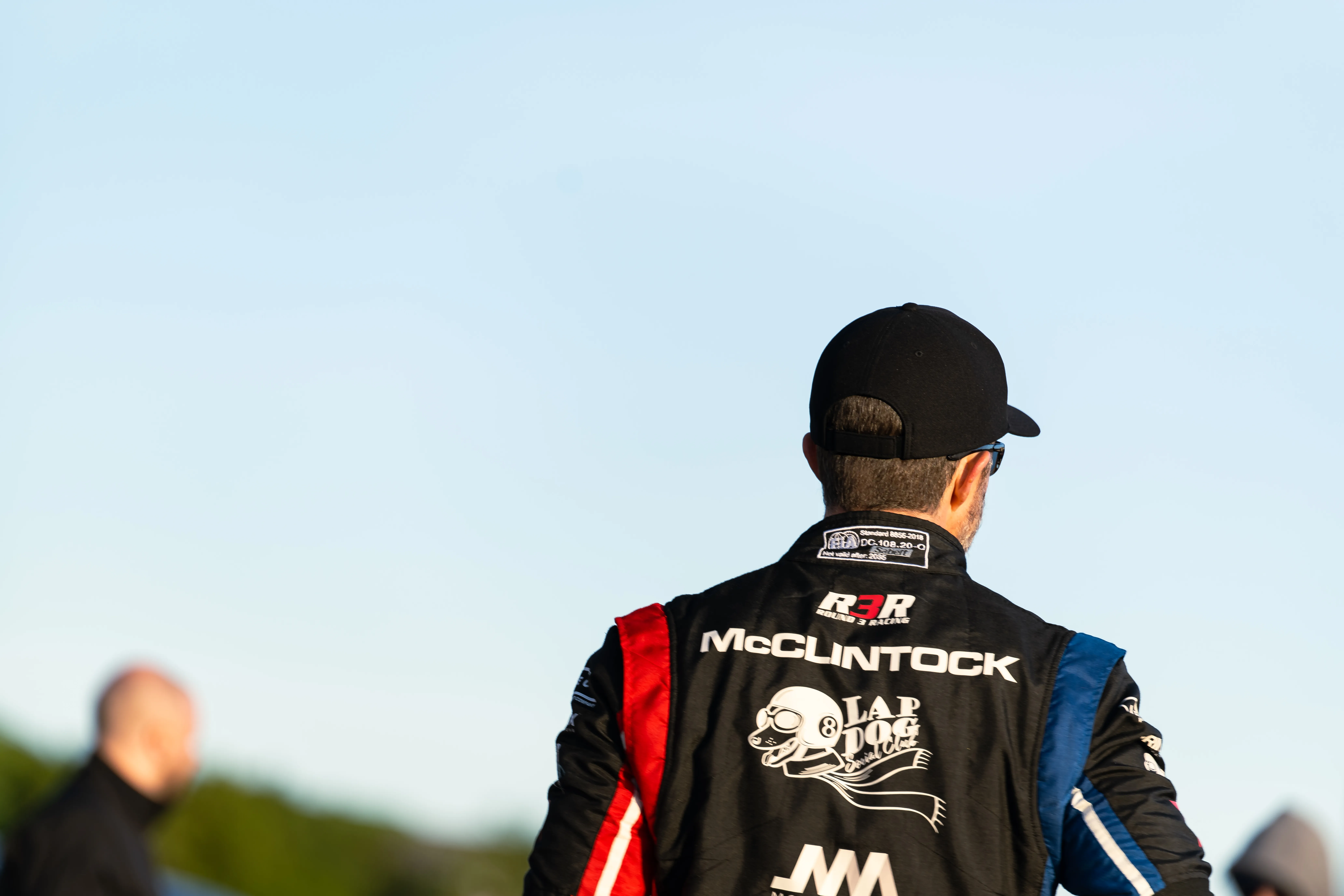 Jon McClintock from behind in his race suit, his name across the shoulders, a black cap, the track ahead at dusk.