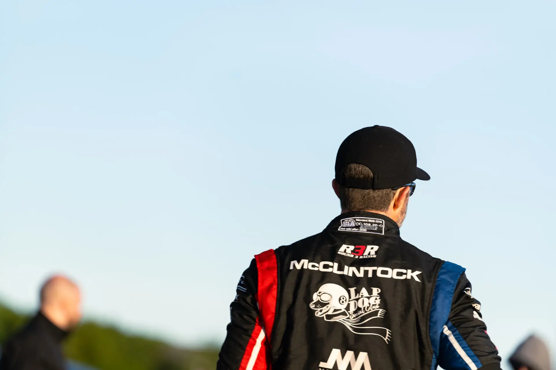 Jon McClintock from behind in his race suit, his name across the shoulders, a black cap, the track ahead at dusk.