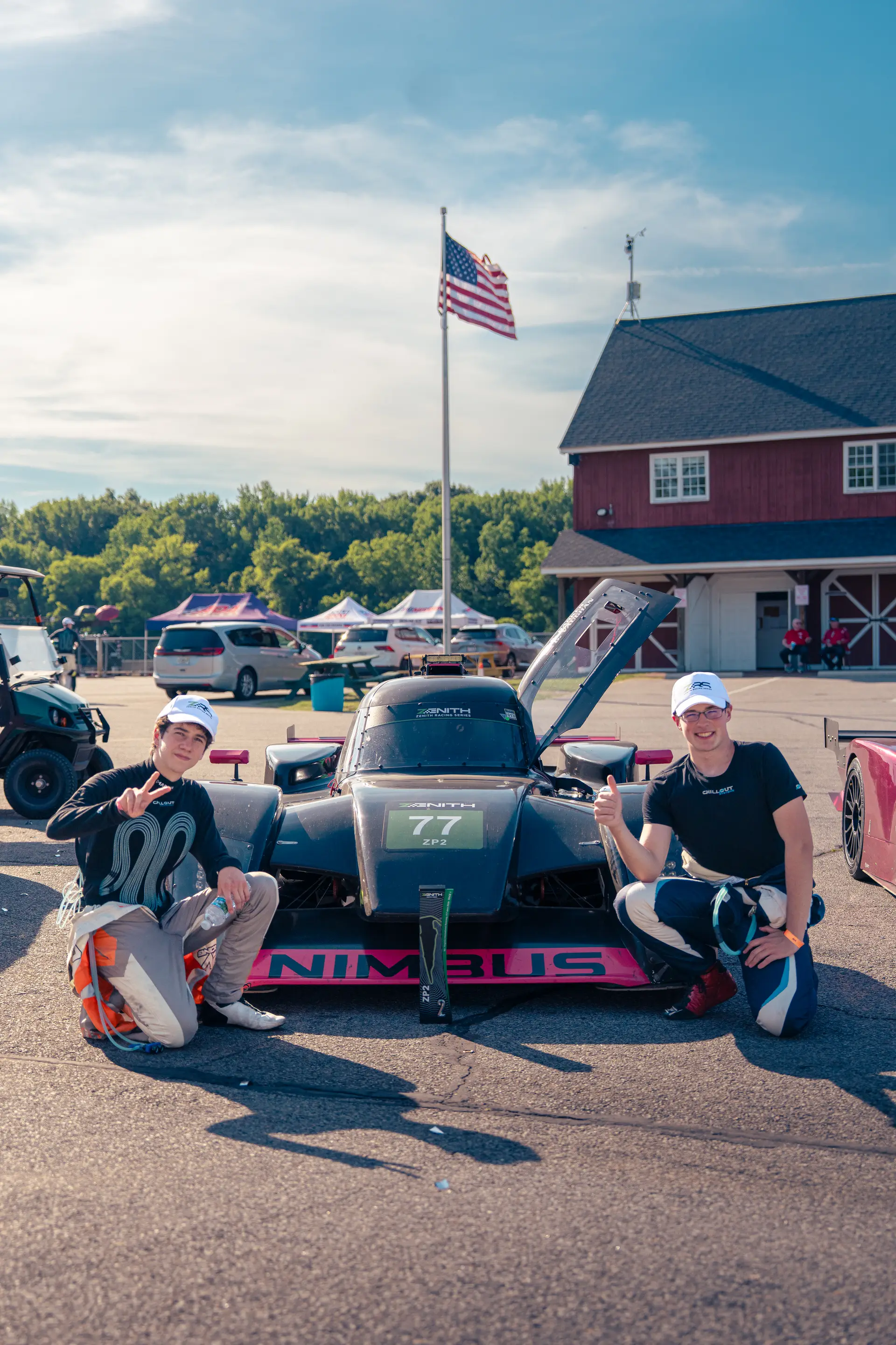 Gavin Vaughan and Ethan Barker crouched on either side of the Nimbus #77 after the race, both throwing peace signs, the VIR barn and an American flag behind them in golden light.