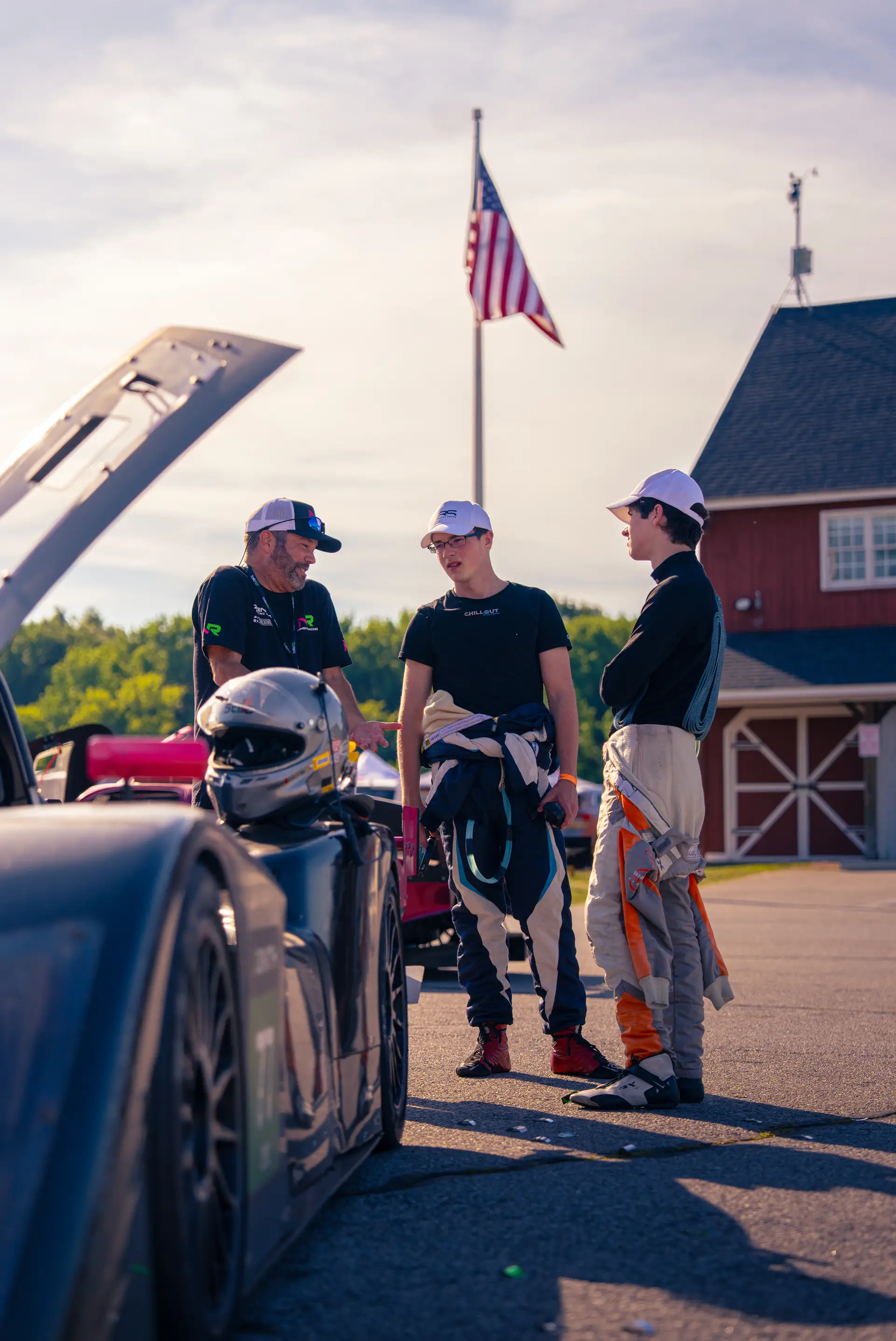 Gavin Vaughan with members of his team in the VIR paddock, the car's bodywork raised beside them, an American flag and the circuit's red barn behind.