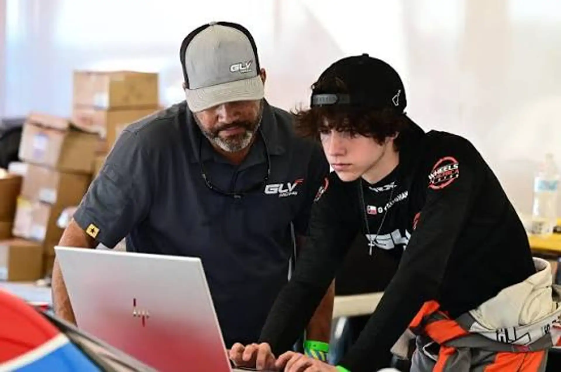 Gavin Vaughan, a teenager in a dark race suit and cap, leaning over a laptop in the paddock alongside a crew member, both studying the screen.