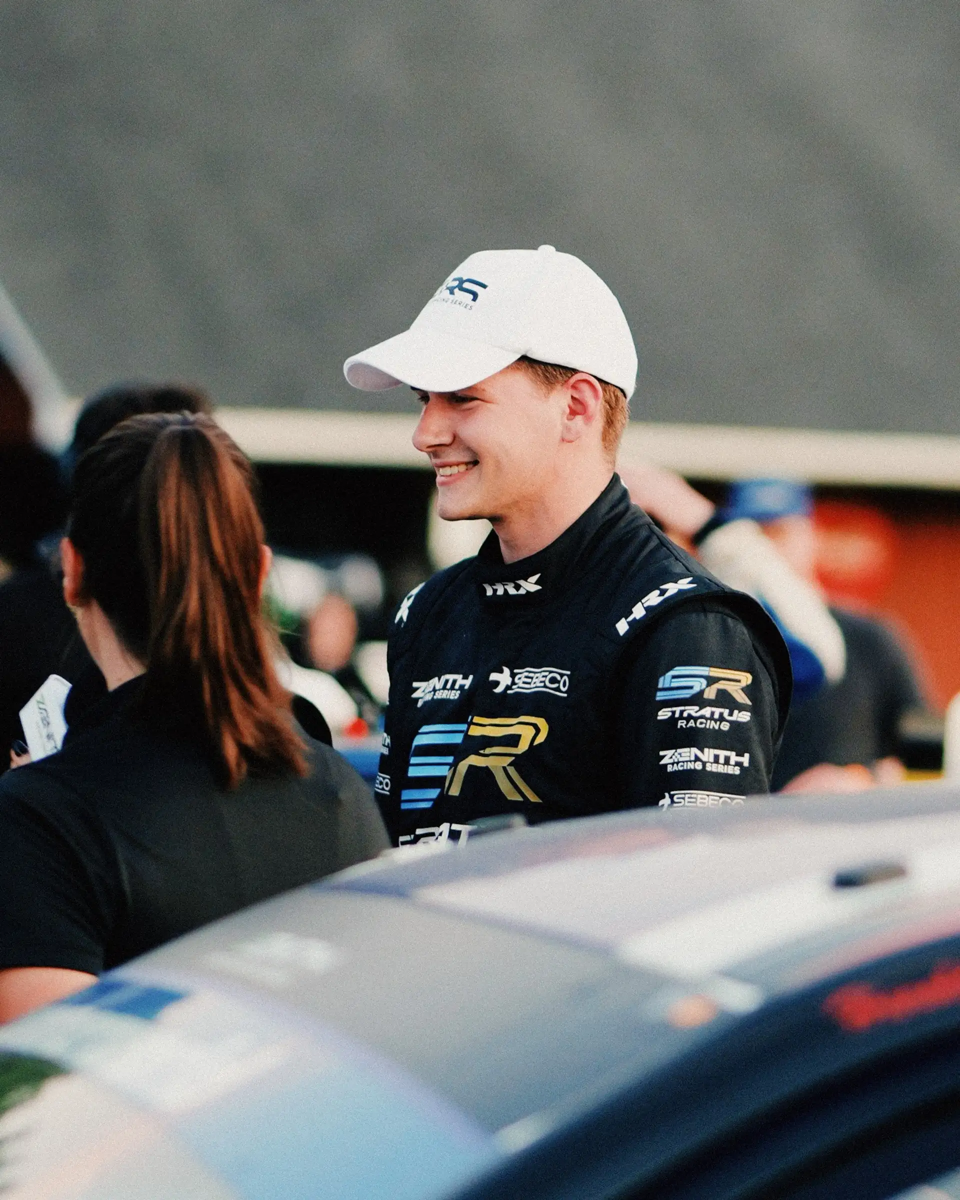 Jason Alder in a white cap and black Stratus Racing driver's suit, smiling in the paddock at golden hour.