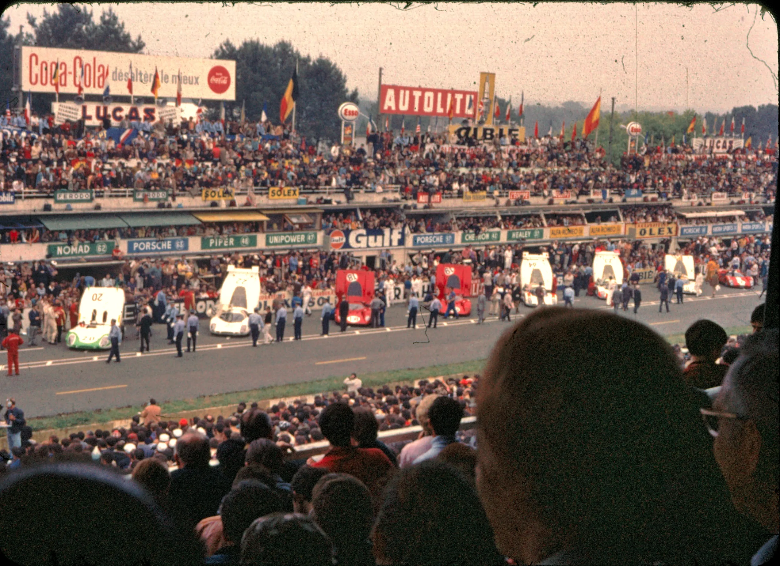 The pre-start grid at the 1969 24 Hours of Le Mans, with Porsche 917s and prototypes lined up against the pit wall, a packed grandstand behind, and period Coca-Cola, Lucas, Gulf, and Autolite signage above the pit boxes.