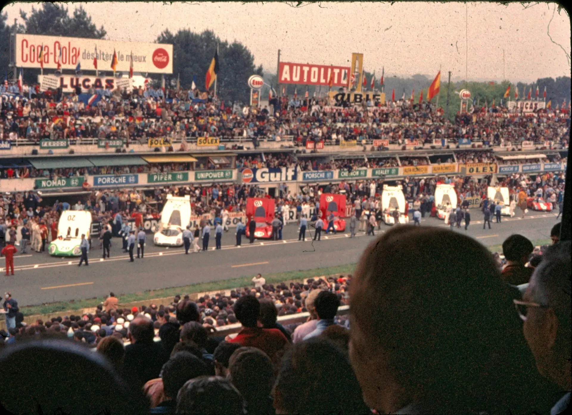 The pre-start grid at the 1969 24 Hours of Le Mans, with Porsche 917s and prototypes lined up against the pit wall, a packed grandstand behind, and period Coca-Cola, Lucas, Gulf, and Autolite signage above the pit boxes.