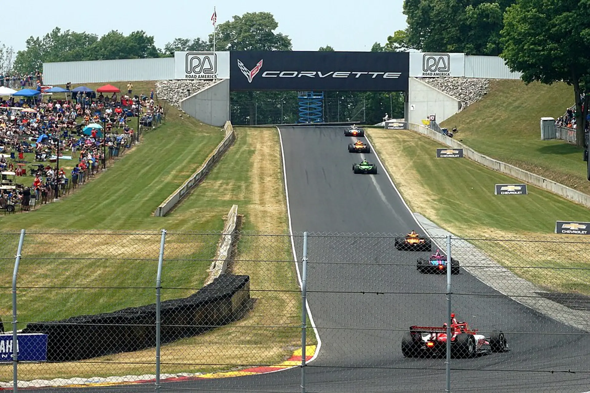 A modern view of Road America: open-wheel cars streaming down one of the circuit's long straights between the trees and the grass banking.