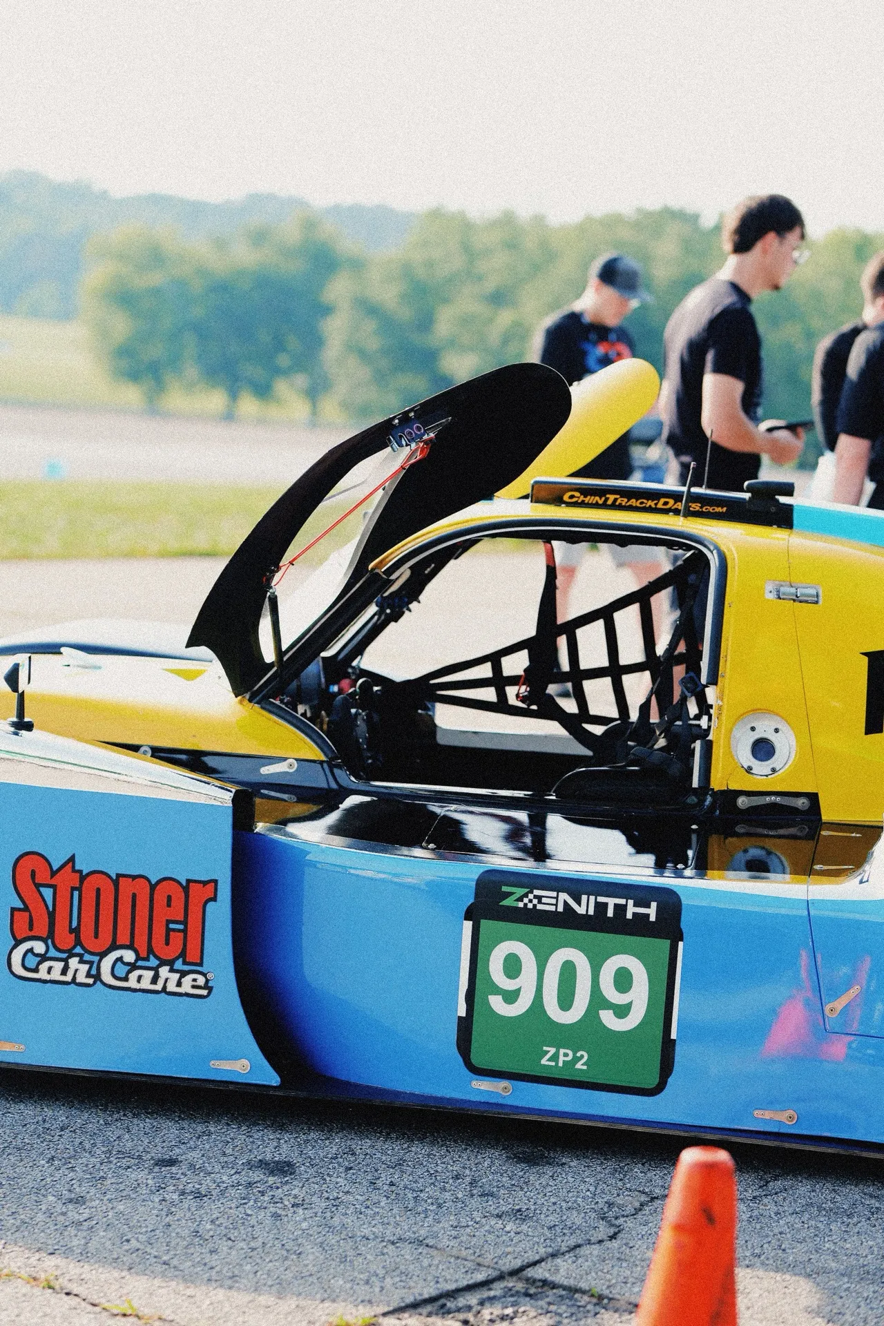 The blue-and-yellow #909 prototype in the garage with its rear bodywork raised, crew standing behind it.