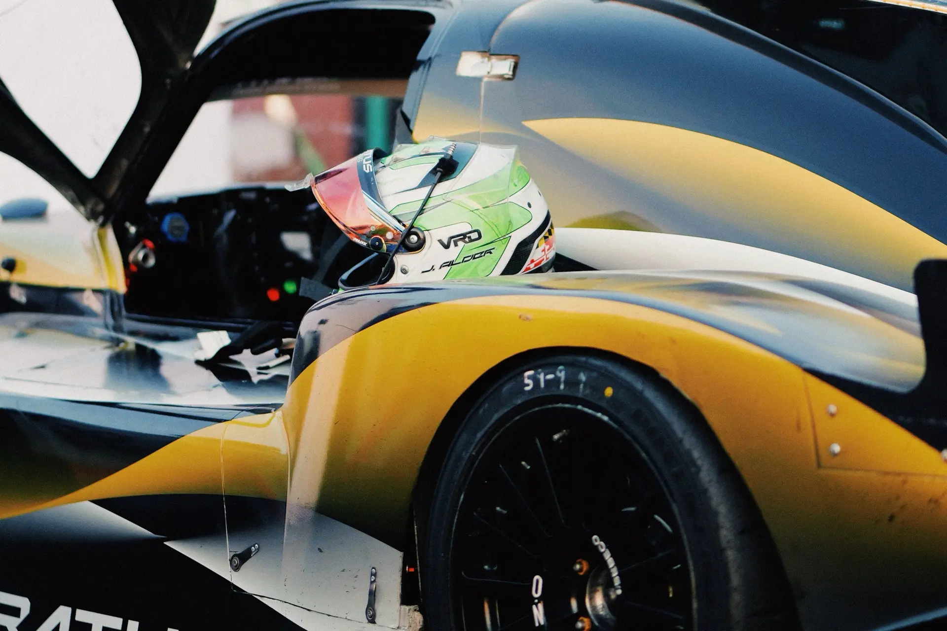 A driver in an iridescent green helmet seated in the open cockpit of the yellow #59 Stratus prototype, engine cover raised, in warm golden-hour light.